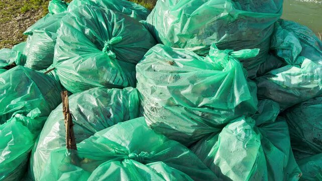 A pile of full green plastic bags sits on the ground near the water. The scene creates a feeling of responsibility mixed with a sadness about pollution. It shows a hardworking effort to clean nature.