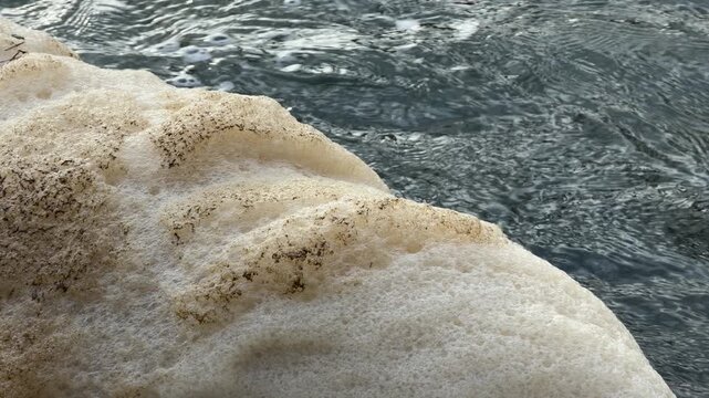 Thick, creamy foam drifts slowly across the dark, rippling water. This heavy accumulation of bubbles creates a curious, soft texture that feels both mysterious and slightly unsettling in nature.