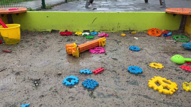 Brightly colored toys lie scattered across wet, muddy sand in an empty playground. The scene feels quiet and bit lonely after a rainstorm. It captures a nostalgic moment of childhood play left behind