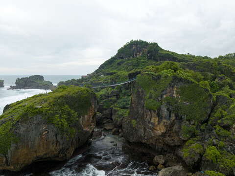 Aerial view of the suspension bridge at Siung Beach connecting rocky cliffs covered in lush greenery with ocean waves crashing below in Tepus, Special Region of Yogyakarta, Indonesia.