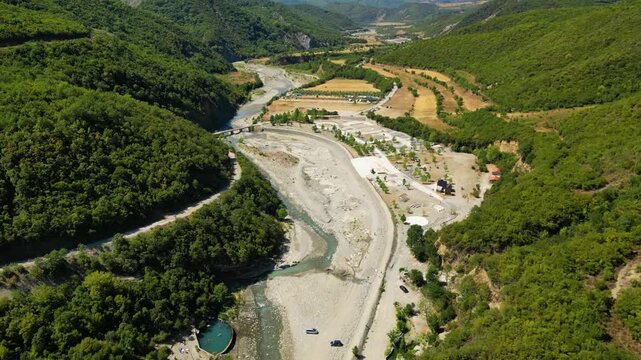 Aerial view of the historic Ottoman Katiu Bridge and circular turquoise thermal baths at the entrance of Langarica Canyon near Permet, Albania, set against the backdrop of the rugged Nemercka