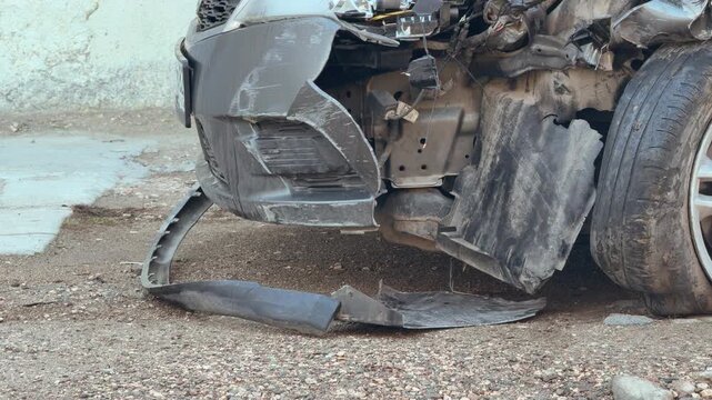 A crashed car shows a detached front bumper resting on the dirt. The sight of the shattered parts and exposed frame feels lonely and unfortunate. This image captures the cold reality of road accident.
