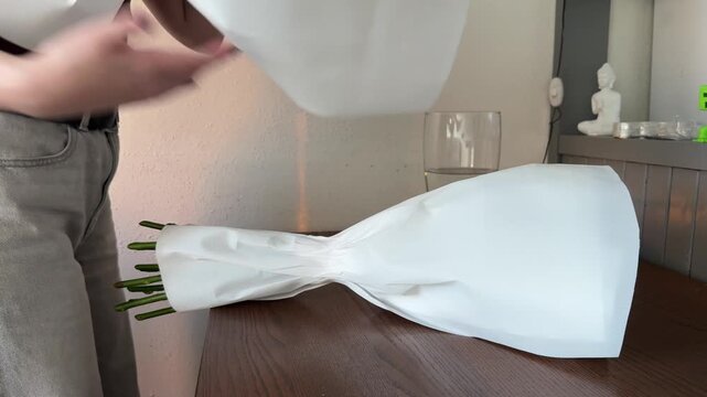 Female person gently untying a recently received flower package, carefully removing its protective paper layer, preparing a fresh bloom for display on a wooden surface.