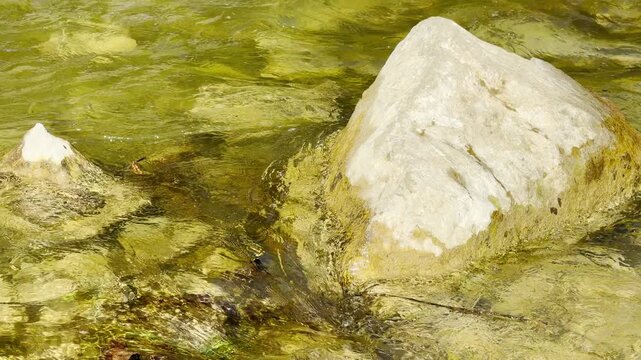 Large, solid rocks sit firmly within a rushing stream of clear, golden water. The shimmering current creates a peaceful and steady feeling of natural strength.