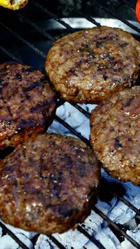 Vertical video: Coals lighting four beef patties, sizzling, charring to sear and brown, cam zooming