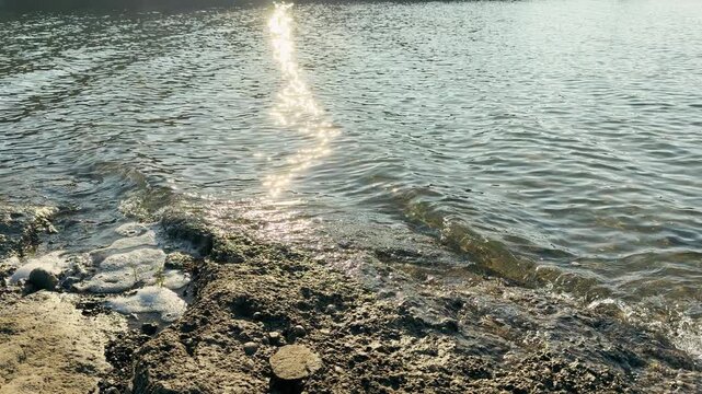 Shimmering sunlight dances on the surface of a quiet lake by a mossy stone bank. Gentle waves wash over the rough earth, creating soft white foam. This scene feels warm, hopeful, and deeply peaceful.