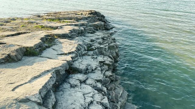 Layered grey rocks stretch out and dip into the shimmering turquoise water. Small patches of grass add life to the rugged stone. The scene feels peaceful, steady, and full of natural wonder.