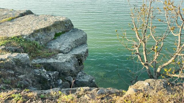 A weathered stone ledge hangs over the calm green water of a peaceful lake. This rugged viewpoint feels sturdy and quiet. The gentle ripples and budding branches create a hopeful, serene mood.