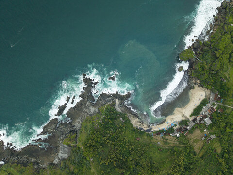 Aerial view of Siung Beach featuring turquoise ocean waves crashing against dark rocky cliffs and a sandy shoreline with lush green vegetation in Tepus, Special Region of Yogyakarta, Indonesia.