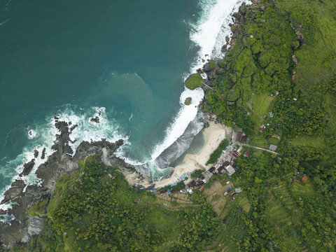 Aerial view of Siung Beach with turquoise ocean waves crashing against jagged rocks, sandy shoreline, and lush green tropical cliffs in Tepus, Special Region of Yogyakarta, Indonesia.