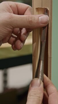 Close-up vertical shot of a craftsman using a chisel to refine a mortise slot in a
wooden door frame. The hand carefully guides the tool to clean and shape the recess, removing small wood shavings.