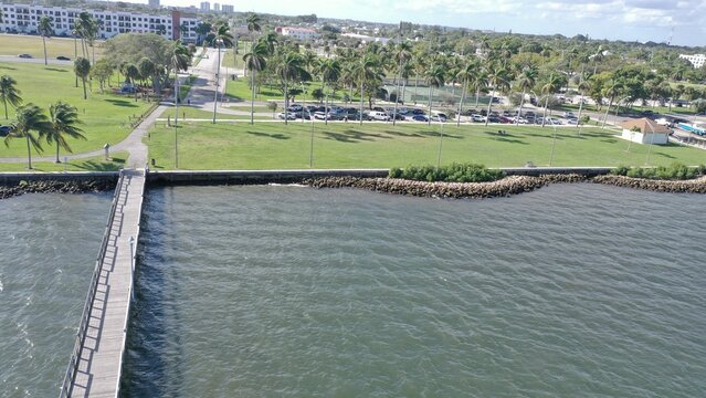 Aerial view of a long wooden pier extending into the sea from a green waterfront park with palm trees and a parking lot.