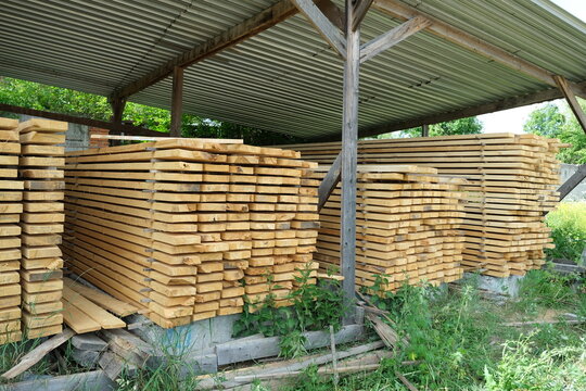 Wooden boards arranged in neat rows with spacers for air drying. Raw timber material stacked under metal roof. Stack of industrial pine wooden planks in an outdoor storage.