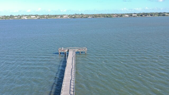 Aerial view of a long wooden pier with people fishing over calm blue water and a distant shoreline with houses and lush trees.