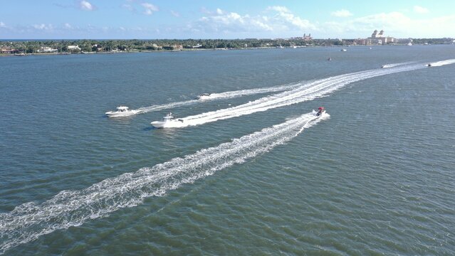 Aerial view of motorboats cruising on the blue water with white wakes near a coastal shoreline with houses and palm trees.