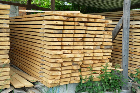 Wooden boards arranged in neat rows with spacers for air drying. Raw timber material stacked under metal roof at construction site or carpentry workshop.