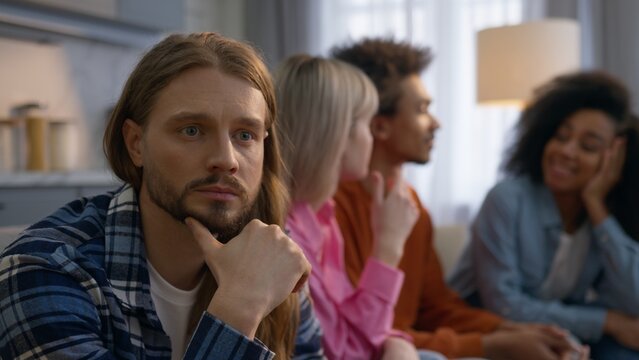 Sad pensive Caucasian man think alone home frustrated introvert feeling separated ignoring friends outsider male while African multiethnic women men laugh talk together friendship antisocial problem