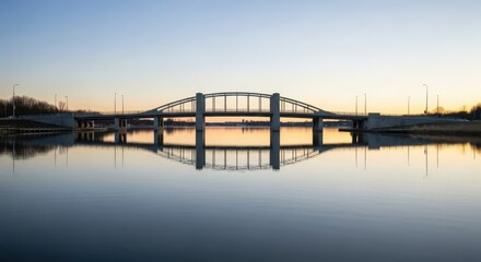 Obraz premium A majestic arch bridge reflects on calm water during a vibrant sunset with clear sky