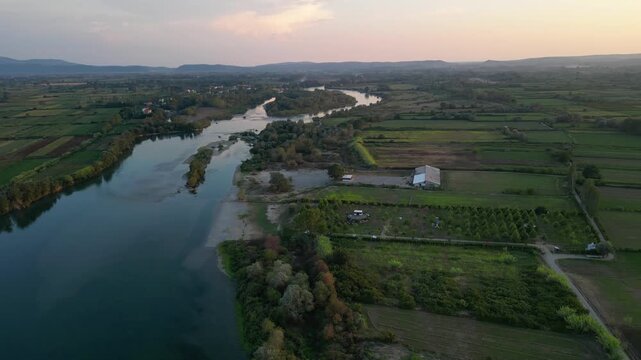 Aerial panorama of a small, green eco-campsite nestled along the banks of the Buna River in Northern Albania, featuring lush agricultural fields and mountain views under a soft sunset sky.
