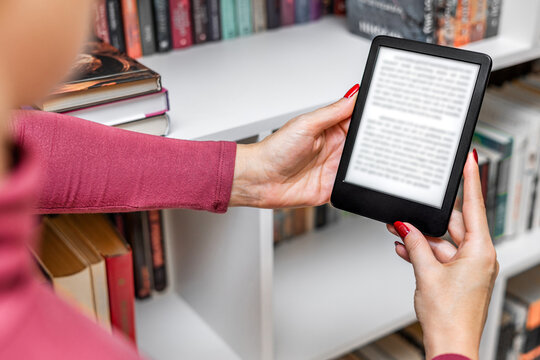 a woman holds an e-book in her hand. A woman reads an e-reader near a bookcase. an e-book against the background of paper books. ease of use of an e-book. a modern way to read books