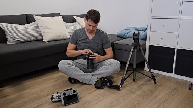 Man Sits Cross-legged On Wooden Floor, Inspecting Mirrorless Camera - Preparing Filming Equipment. wide shot