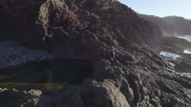 Adventurous young woman standing on a rugged volcanic coastline, enjoying the scenic ocean view next to a natural rock pool
