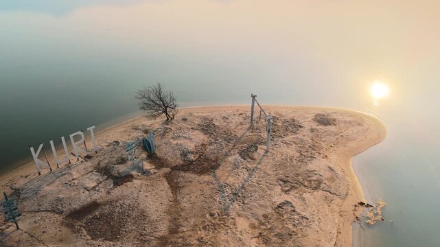 Sandy peninsula extending into calm water, showcasing large 'kurki' letters, an old tree, and a wooden swing set, all basking in the warm light of a late afternoon sun or early morning sunrise
