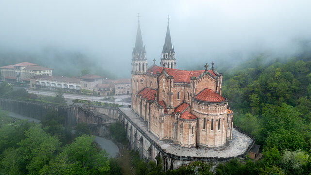 Aerial view of the magnificent Basilica de Santa Maria la Real de Covadonga emerges through the mist, its spires piercing the sky above lush greenery, Cangas de Onis, Spain.