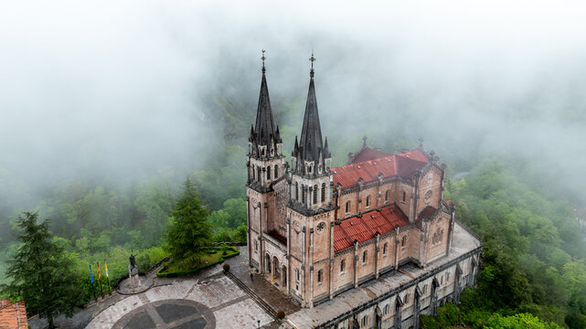 Aerial view of the Basilica's spires piercing through the dense fog, its red-tiled roof contrasting with the lush green forest, Basilica de Santa Maria la Real de Covadonga, Cangas de Onis, Spain.