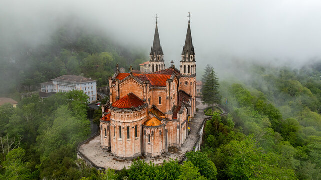Aerial view of Basilica piercing through the misty landscape, its spires reaching towards the heavens above the lush greenery, Basilica de Santa Maria la Real de Covadonga, Cangas de Onis, Spain.
