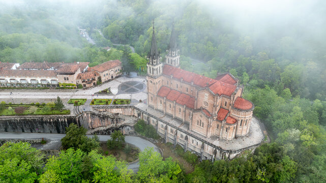 Aerial view of the Basilica de Santa Maria la Real de Covadonga emerges through the mist-laden forest, a tapestry of red roofs and stone spires, Cangas de Onis, Spain.