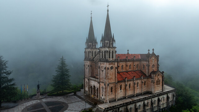 Aerial view of the Basilica's spires pierce the misty veil, its aged stone glowing softly amidst the lush, green landscape, Basilica de Santa Maria la Real de Covadonga, Cangas de Onis, Spain.