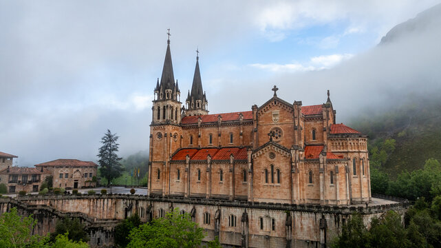 Aerial view of the Basilica de Santa Maria la Real de Covadonga stands majestically against the misty mountain backdrop, its red roofs contrasting with the stone facade, Cangas de Onis, Spain.