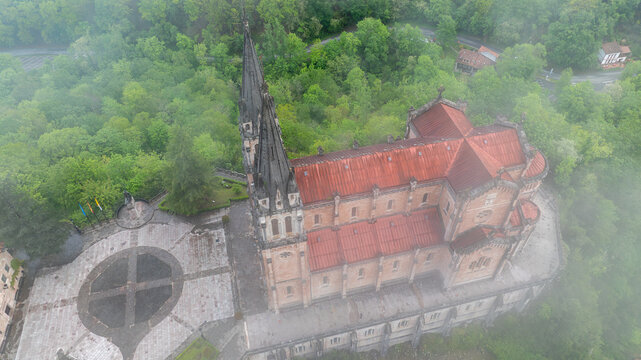 Aerial view of the Basilica's red roofs contrast with the surrounding green forest, shrouded in mist, Basilica de Santa Maria la Real de Covadonga, Cangas de Onis, Spain.