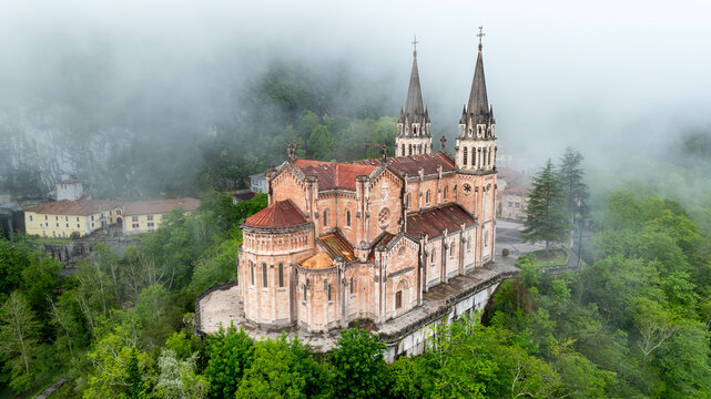Aerial view of the Santa Maria la Real de Covadonga Basilica, its spires piercing through the mist-shrouded mountains, a contrast to the lush green forests below, Cangas de Onis, Spain.