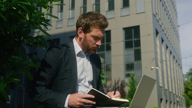 Angry stressed Caucasian business man working laptop sit outside city multitasking male annoyed businessman guy employer online typing computer writing notes documents mistake bad result loss downtown