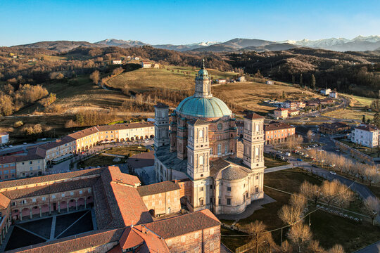 Aerial view of the Santuario di Vicoforte featuring its massive elliptical dome and historic complex surrounded by rolling hills and the snow-capped Alps Vicoforte, Piedmont, Italy.