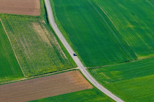 Aerial view of vibrant emerald green and rich earthy brown fields dissected by a winding grey road with a lone car, Wintzenheim-Kochersberg, Grand Est, France.