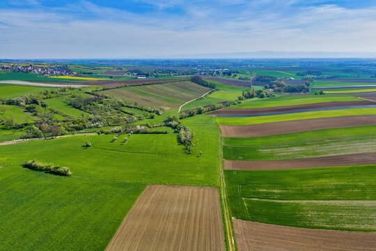 Aerial view of a vibrant tapestry of green and brown fields across rolling hills, a distant village visible, Wintzenheim-Kochersberg, Grand Est, France.