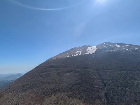   
Paesaggio montano con pendii boscosi in ombra e una vetta pi&ugrave; alta illuminata dal sole, con tracce di neve e cielo limpido. Un&rsquo;atmosfera luminosa e naturale