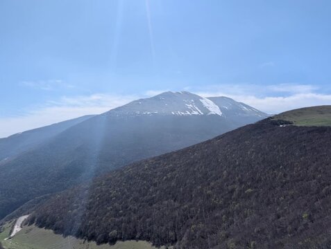   
Paesaggio montano con pendii boscosi in ombra e una vetta pi&ugrave; alta illuminata dal sole, con tracce di neve e cielo limpido. Un&rsquo;atmosfera luminosa e naturale