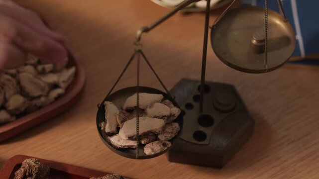 Traditional herbalist weighs dried root slices on a small balance scale. Focus on precision, dosage control and authentic apothecary process in oriental medicine preparation.