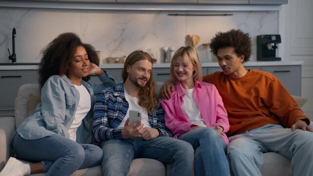 Multiethnic students friends men and women looking at mobile phone at home couch together Caucasian girls African American guys talking chatting news social media sharing diversity friendship indoors