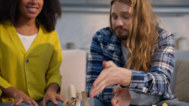 Multiethnic couple playing jenga game at home Caucasian man African American woman stacking wooden blocks tower fun laughing joy playful boyfriend girlfriend husband wife girl accidentally falls smile
