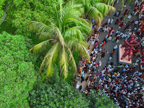 Dhaka, Bangladesh - 14 April 2026: Aerial view of a vibrant crowd of devotees gathered around a towering, reddish deity statue, contrasting sharply with the lush, verdant canopy of tropical trees.