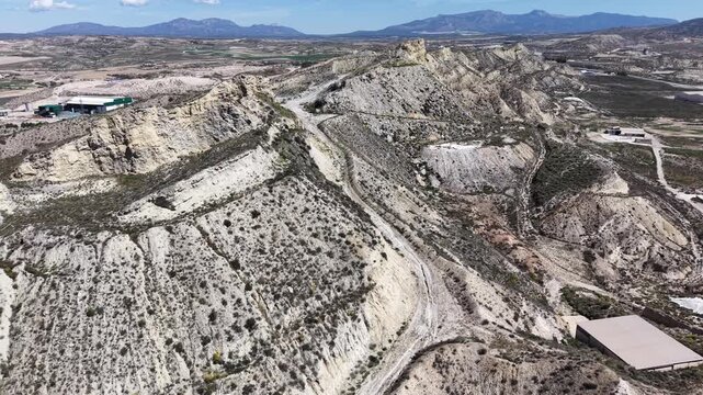 A tracking drone shot moving across the arid, eroded badlands of Lorca, showing steep ridges and a water deposit near industrial sites.