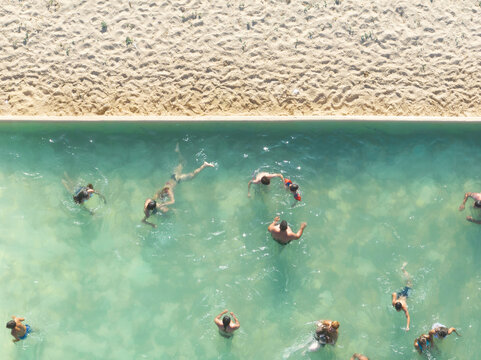 Figueira da Foz, Portugal - 08 August 2024: Aerial view of swimmers enjoying the refreshing turquoise waters of a pool, sharply contrasted by the golden textured sand under the bright summer sun.
