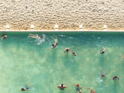 Figueira da Foz, Portugal - 08 August 2024: Aerial view of a public swimming pool, where figures splash in clear turquoise waters beside a sun-drenched sandy beach.
