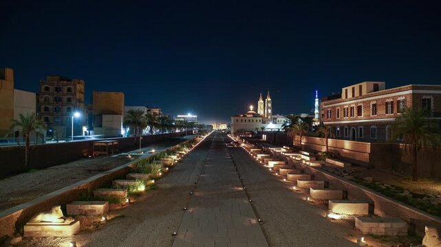 Night time lapse of illuminated Avenue of Sphinxes leading toward Luxor temple in Egypt, flanked by palms, historic architecture ruins and church under a clear starry sky.