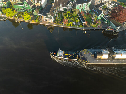 Zaanse Schans, Netherlands - 29 April 2025: Aerial view of a tugboat and barge on the dark canal, framed by vibrant green traditional Zaanse Schans houses.